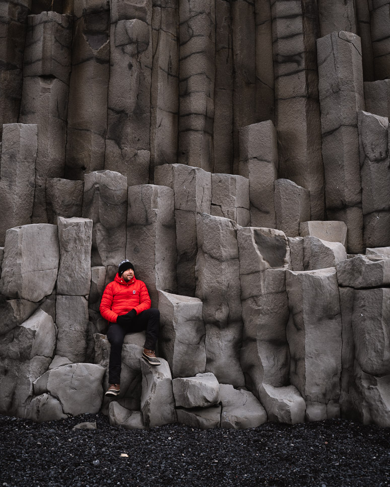 Man met rode winterjas op de grijze basalt zuilen van het Reynisfjara zwarte strand, langs de ring road  in Zuid IJsland