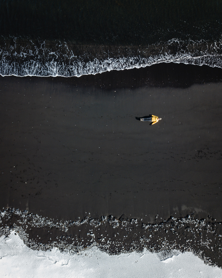 Dronefoto van Hvitserkur strand met een vrouw die ligt op het zwarte lavastrand met boven haar de zee en onder haar de sneeuw