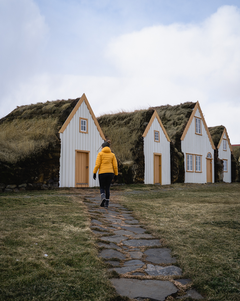 Vrouw in gele jas wandelt naar de turfhuisjes met grasdaken in Glaumbær, een verborgen parel in Noord IJsland
